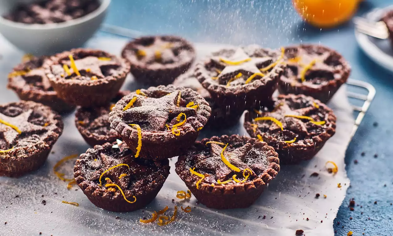 Chocolate-orange mince pies with crumbly brown pastry, golden orange zest, sugar dusting, and star-shaped tops on parchment paper