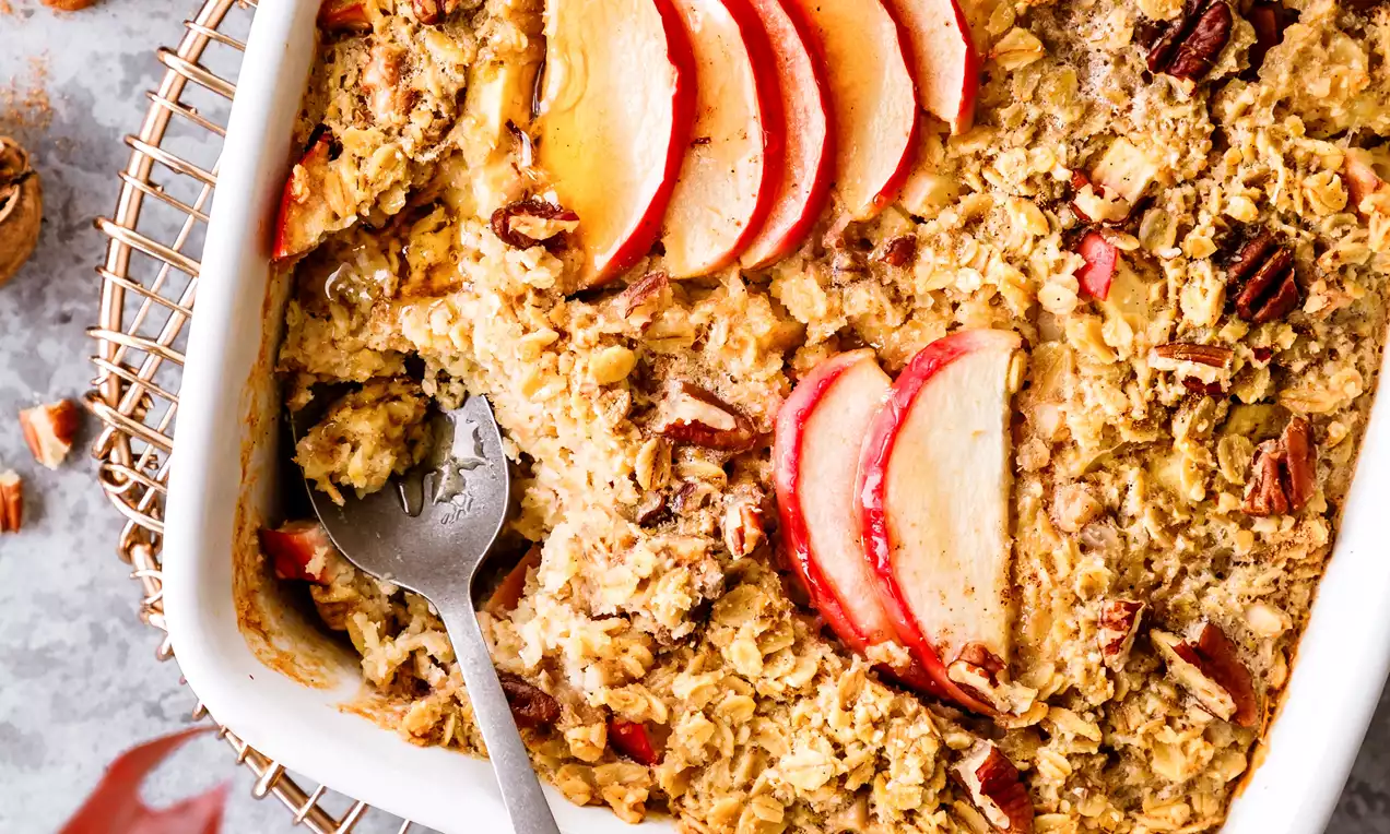 Picture - Baked apple oatmeal in a white square dish with a golden surface, pecan pieces, and sliced red apple arranged on top