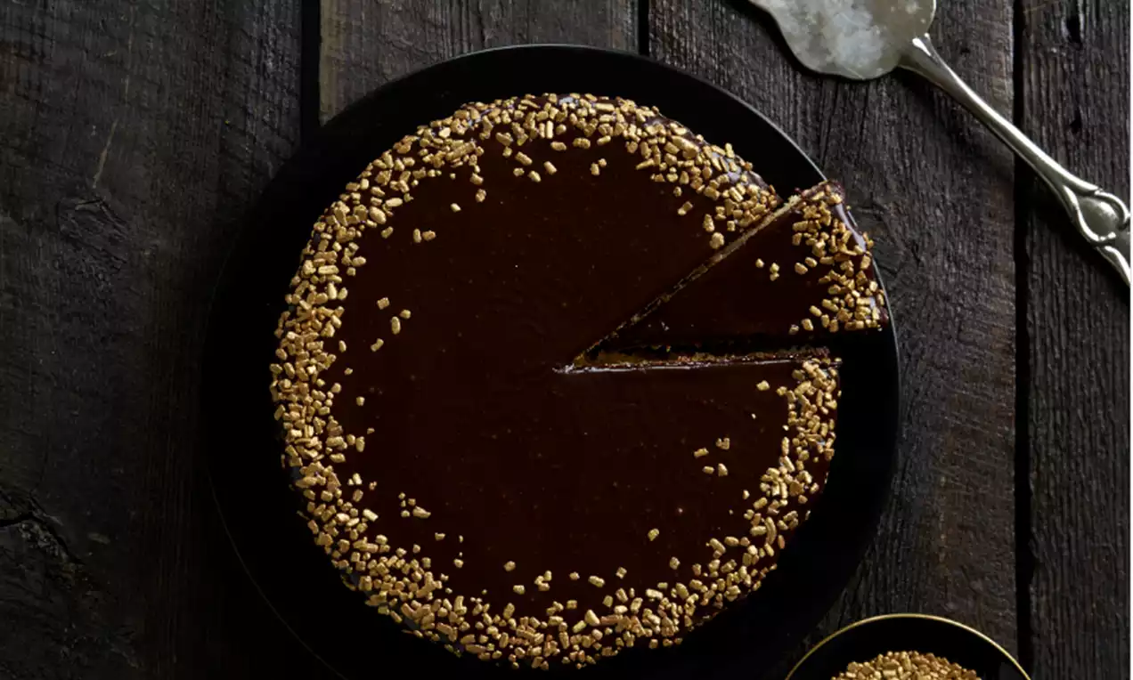 Dunkler Schokokuchen mit glänzender Glasur, goldenen Krokantstückchen am Rand, auf schwarzem Teller, daneben Metalltortenheber und Krokantschale