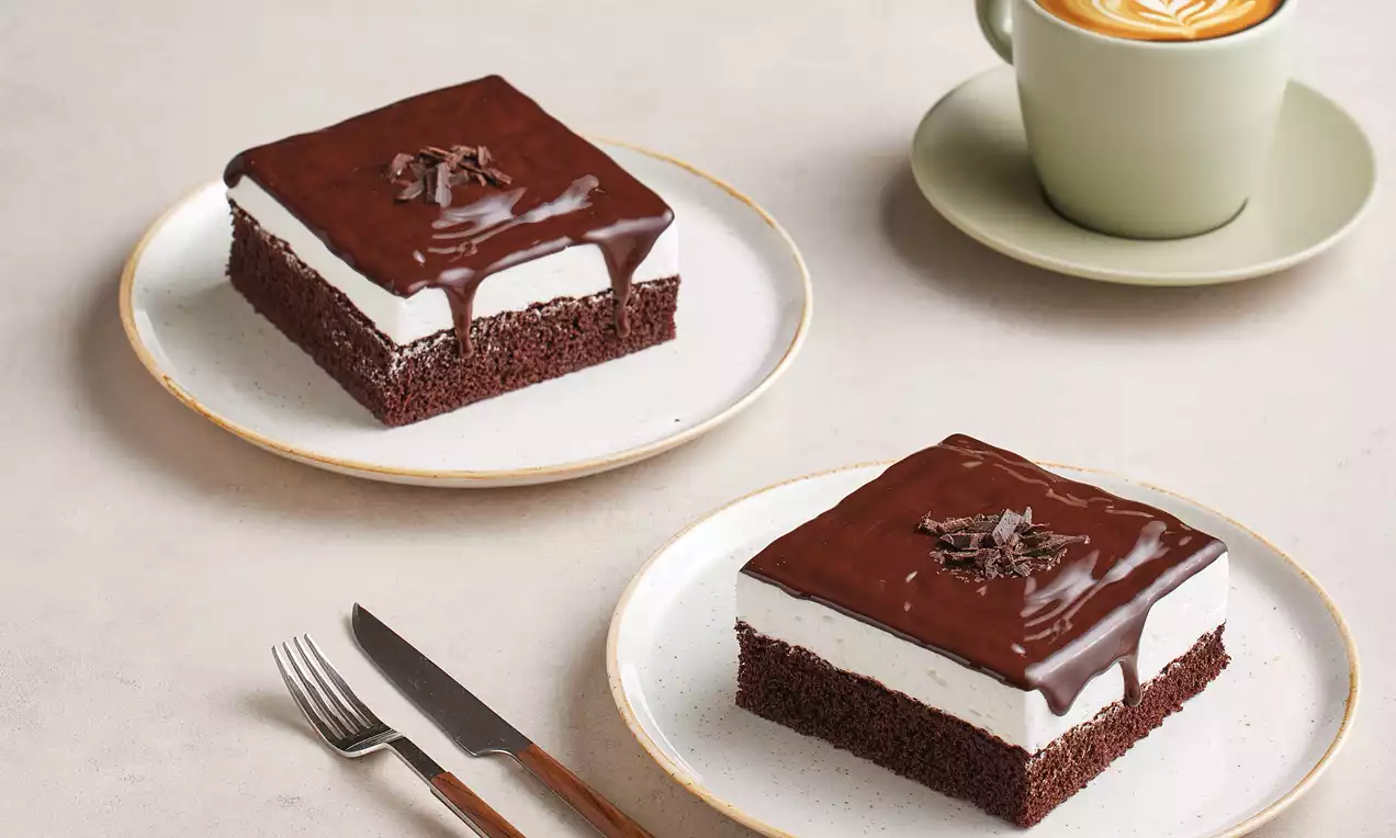 Kakao tabanlı kare dilimler, beyaz krema katmanı, parlak çikolata sos ve çikolata rendesiyle açık renkli tabakta sunulan tatlı