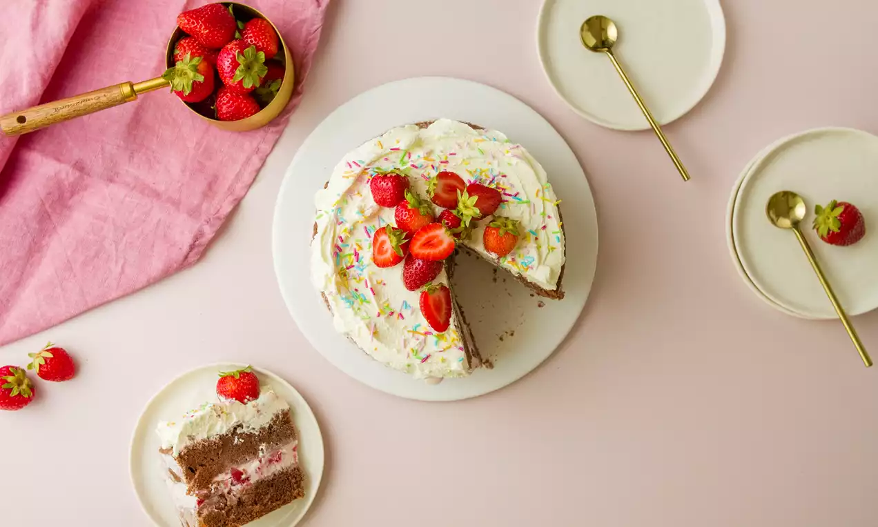 Tarta helada cilíndrica con crema blanca y virutas de colores, decorada con fresas enteras y partidas sobre un plato blanco
