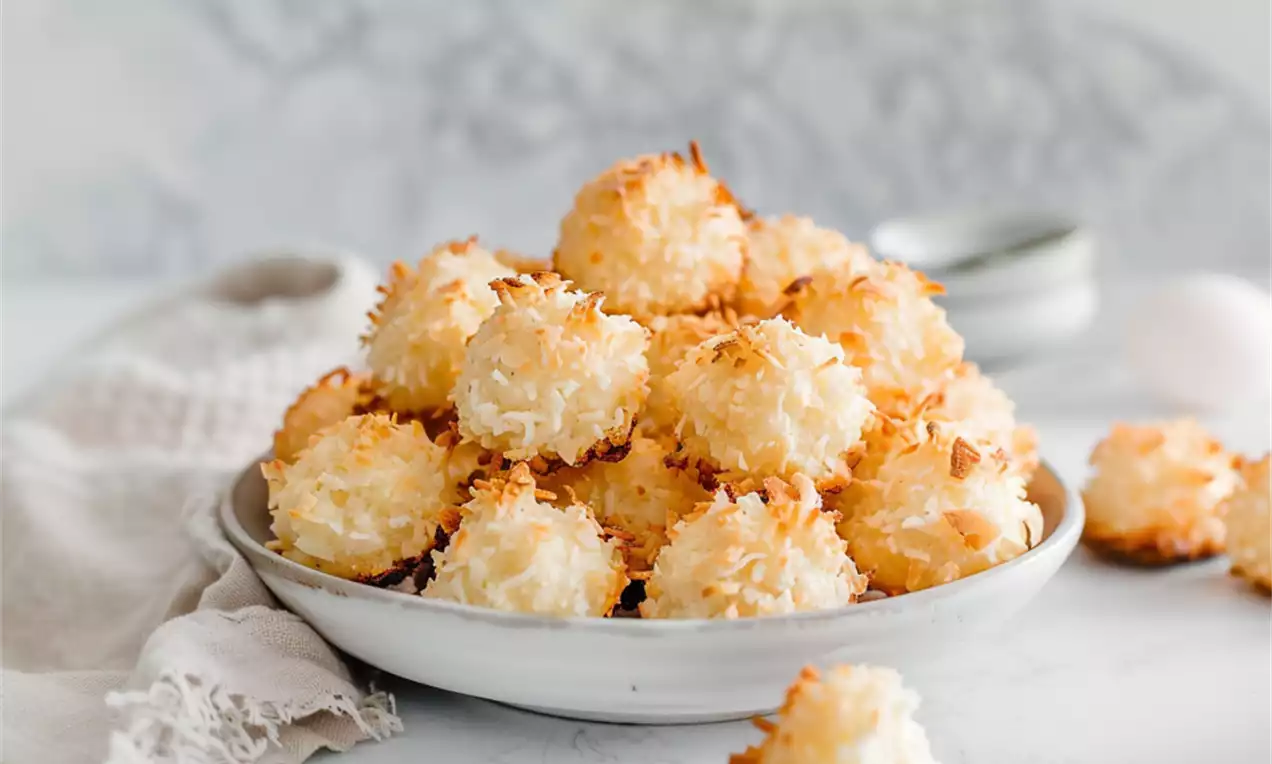 Golden coconut macaroons with toasted edges and rough, flaky texture piled in a white bowl on a table with a linen cloth and blurred jars