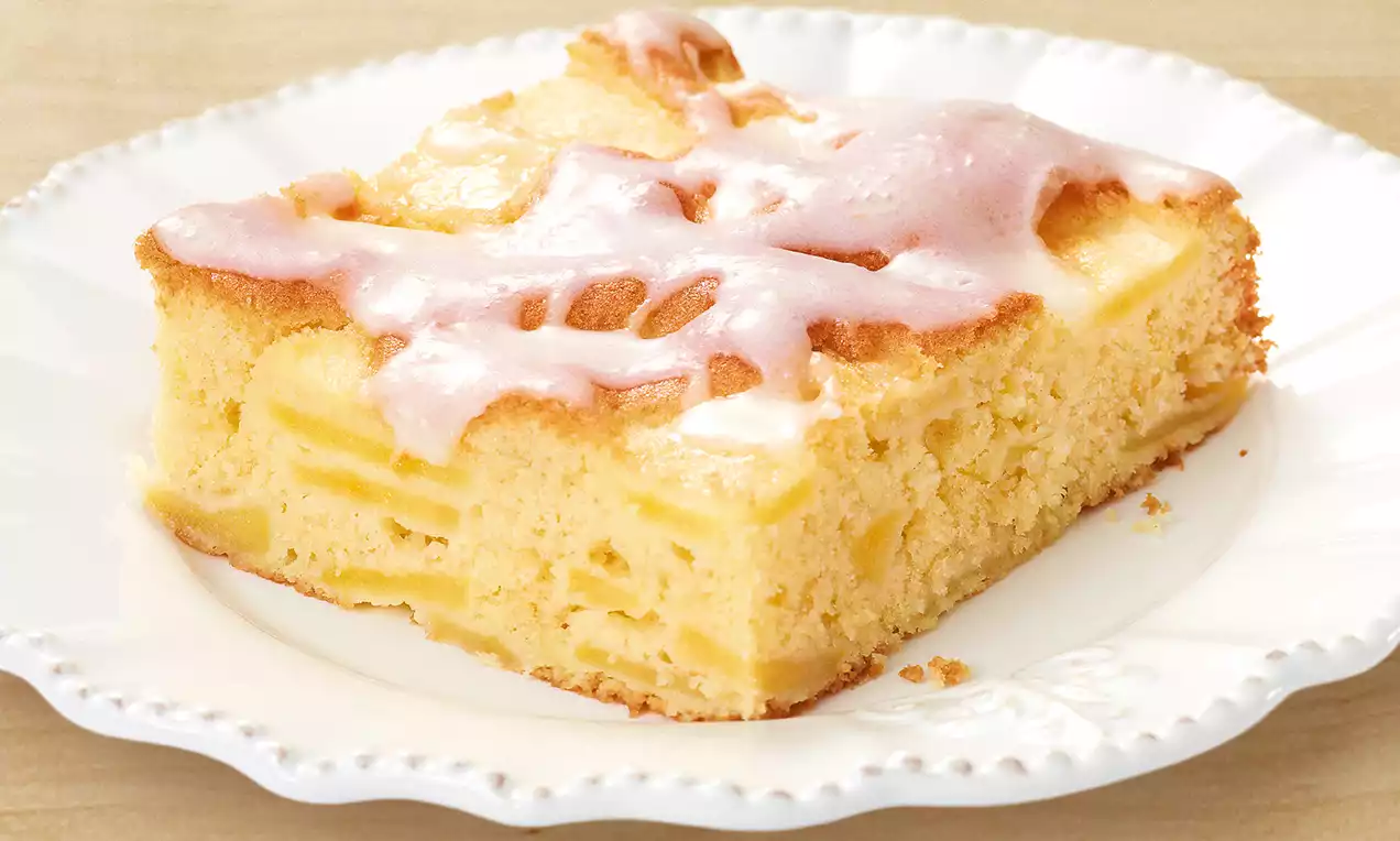 Ein Stück goldgelber Apfelkuchen mit zarter Glasur und sichtbaren Apfelstücken auf einem weißen, gewellten Teller