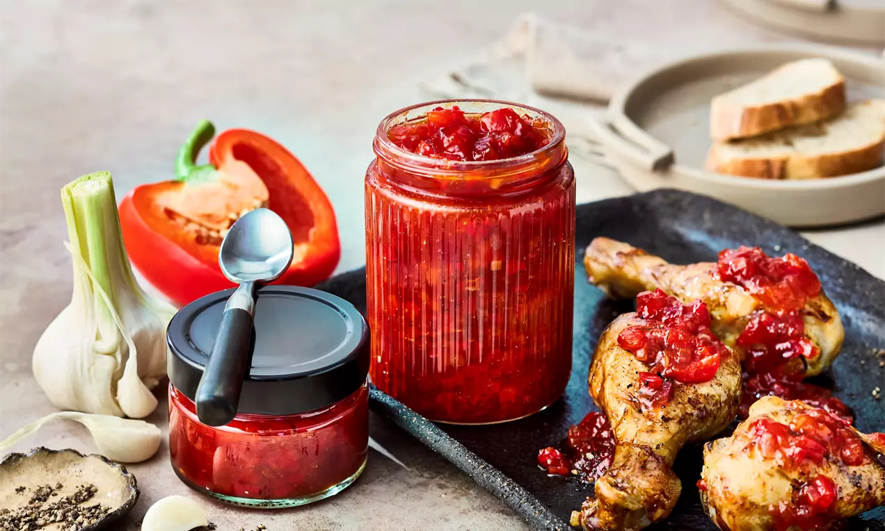 Glänzendes rotes Paprika-Chutney in zwei Gläsern neben gebackenen Hähnchenschenkeln mit Chutney, Paprika und Knoblauch auf rustikalem Tisch