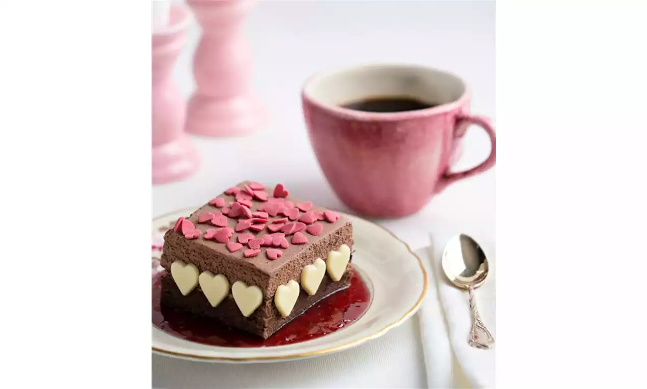 Rektangulär chokladbakelse med vitahjärtformade chokladelement, rosa strössel på toppen, liggande på hallonsås på en vit desserttallrik