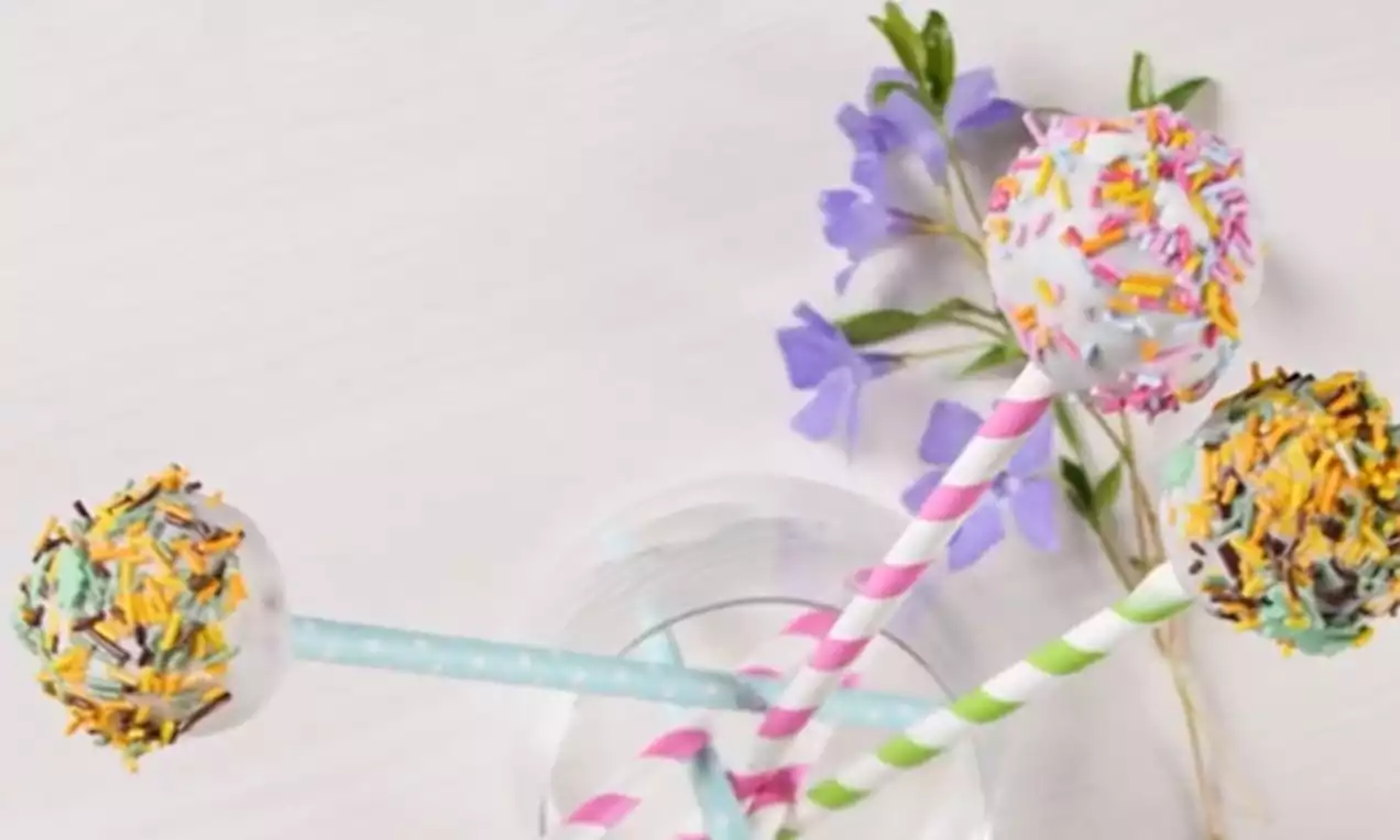 Trīs apaļi kūciņu kēksiņi uz svītrainiem salmiņiem, pārklāti ar balto glazūru un dažādu krāsu cukura dekoriem, uz gaiša fona ar ziedu akcentu