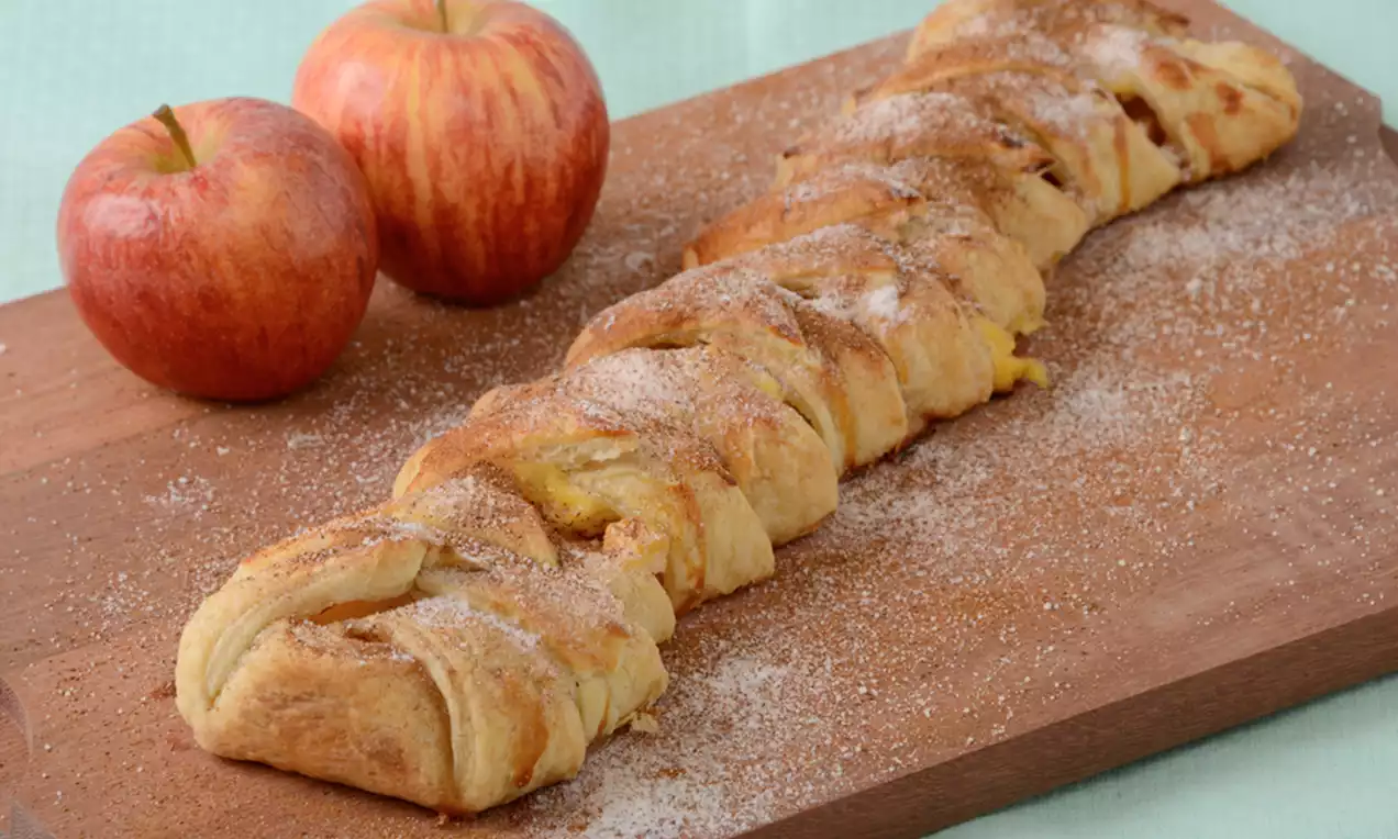 Trança dourada de massa folhada entrelaçada com recheio cremoso, polvilhada com açúcar e canela sobre tábua de madeira, com maçãs vermelhas ao lado