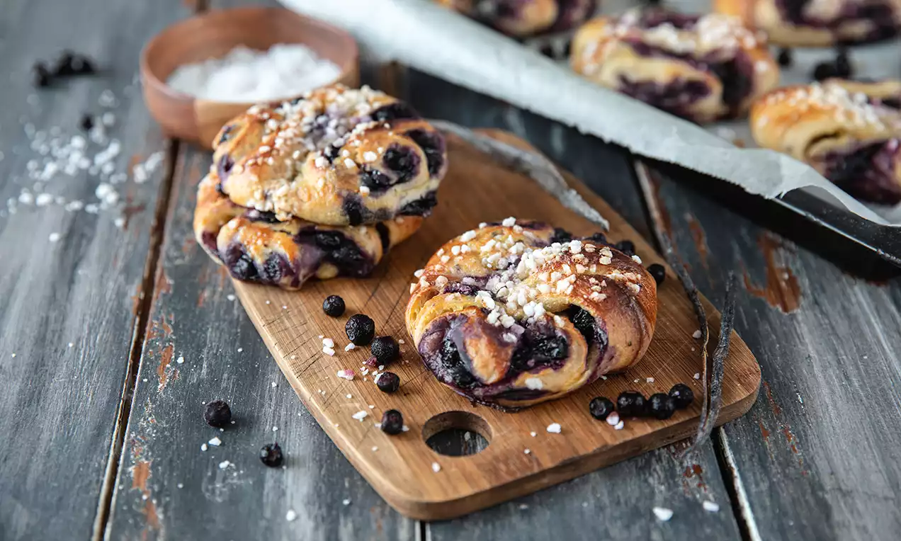Gyldenbrune knuter med blåbær og sukkerperler på en rustikk trefjøl, omgitt av blåbær, en vaniljestang og salt i en skål