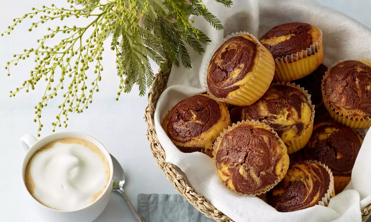 Mramorové muffiny se zlatavou vanilkovou a tmavou kakaovou vrstvou v papírových košíčcích uspořádané v proutěném košíku vedle šálku cappuccina