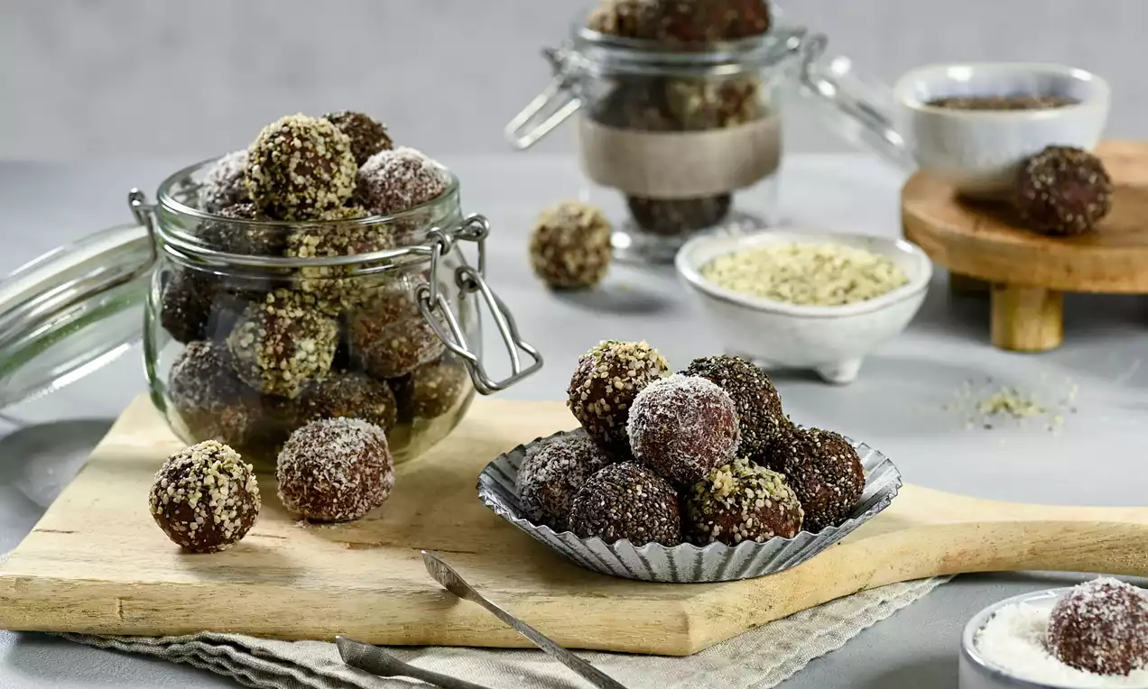 Vegan coconut balls covered in crushed nuts and chocolate, round and textured on a wooden board with glass jars and small bowls nearby
