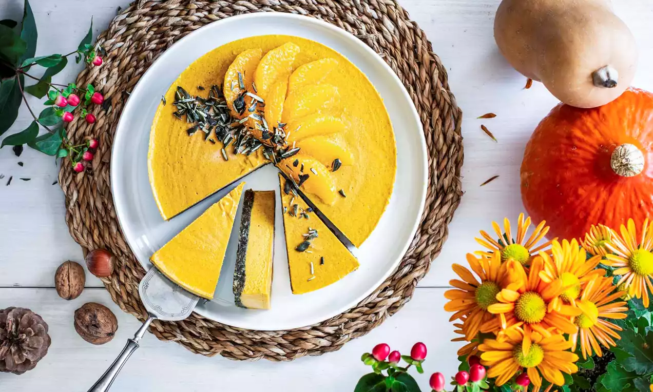 Vegan pompoencheesecake met feloranje vulling, partjes pompoen en zwarte pitjes op een wit bord met bloemendecoratie en herfstgroen