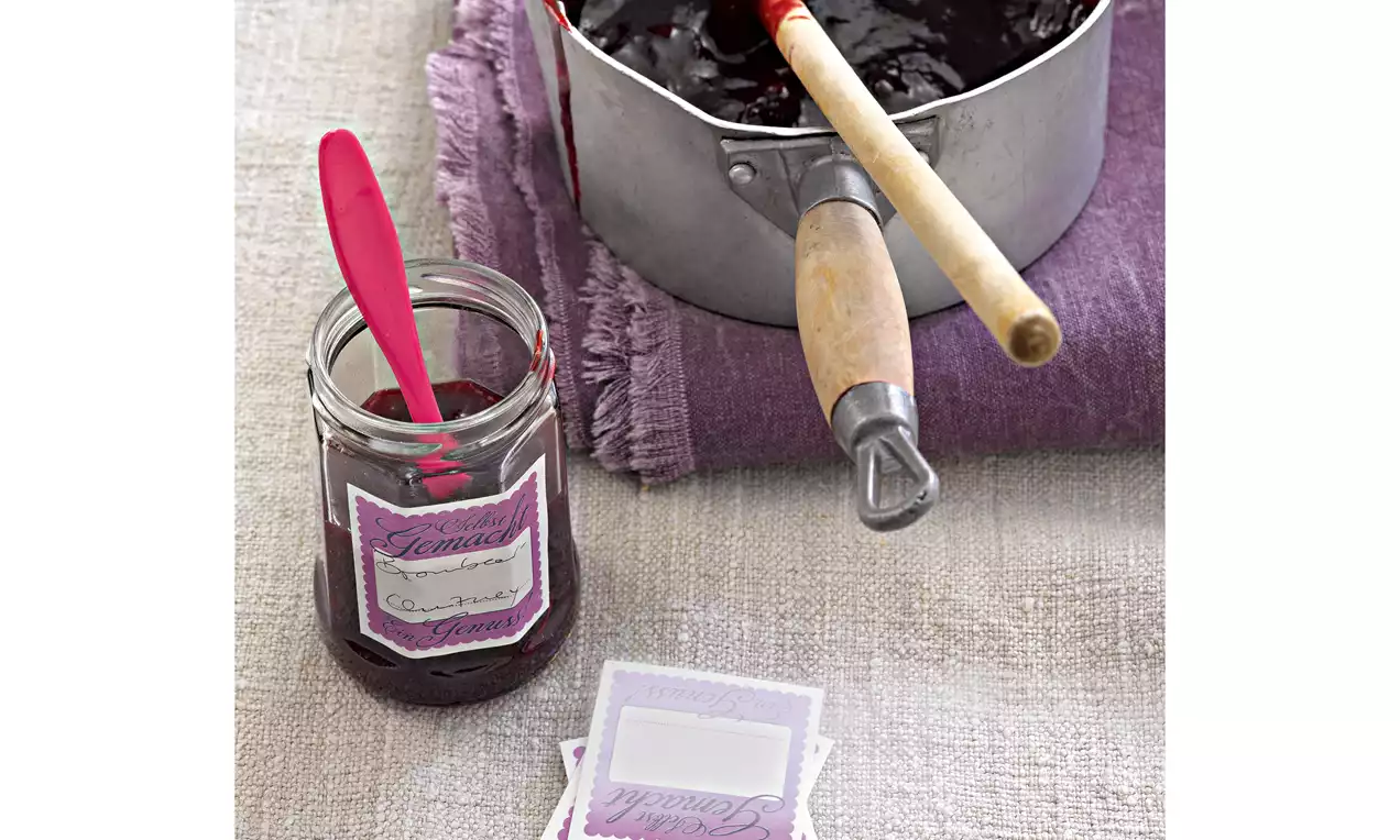 Tiefrote, dickflüssige Brombeer-Schalotten-Masse in einem Topf mit Holzgriff und in einem Glas mit rotem Löffel auf lilafarbener Serviette