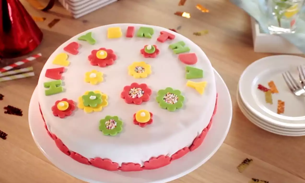 Witte verjaardagstaart met kleurrijke bloemdecoraties en letters in groen, geel en rood op een wit bord, omringd door feestconfetti