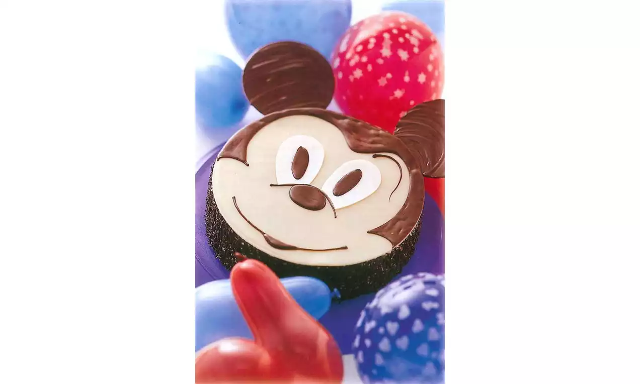 Gâteau rond au glaçage crème représentant le visage de Mickey avec oreilles en chocolat sur une assiette entourée de ballons colorés