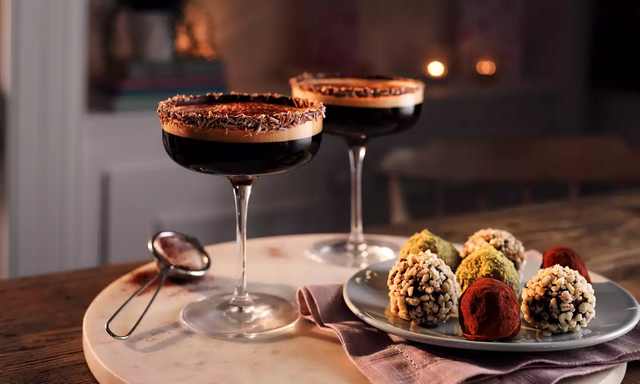 Dark caramel espresso martini with rich coffee foam, chocolate-rimmed glasses, served on a wood table with colourful truffles