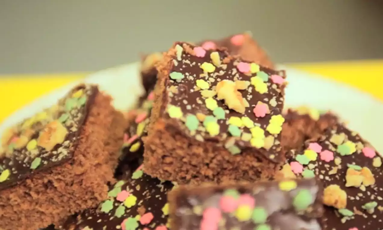 Rutor av chokladkaka med glansig mörk chokladglasyr, strösselpulver i ljusa färger och smulor serverade på en vit tallrik
