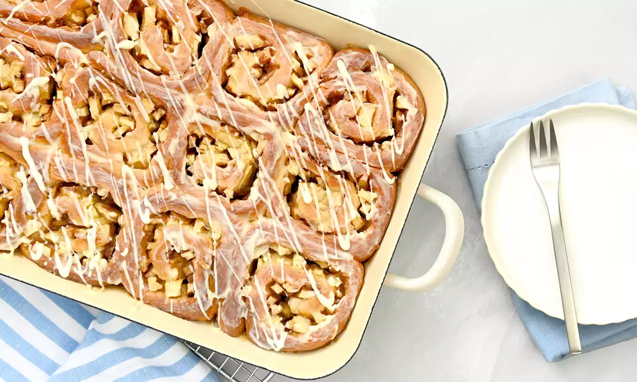 Apfel-Zimtschnecken mit goldbrauner Oberfläche und weißem Zuckerguss in einer hellen Backform, daneben ein Teller mit Gabel auf blauer Serviette