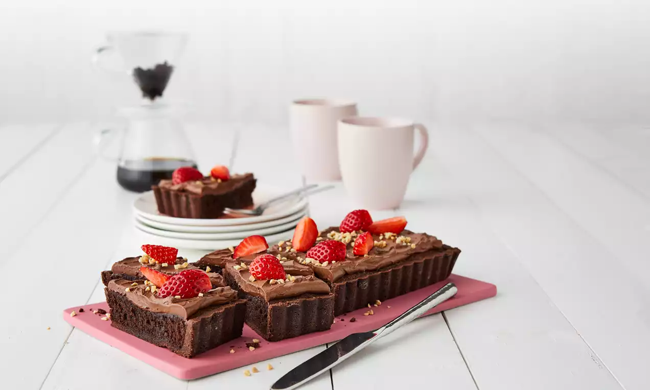 Sjokolademazarin med mørk brun farge, toppet med sjokoladekrem og friske røde jordbær på rosa skjærefjøl med kniv ved siden av