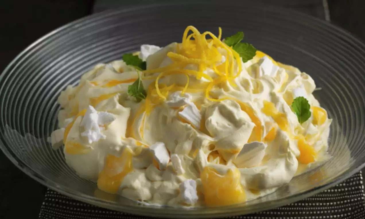 Fluffy pale cream pudding with swirls of yellow lemon curd, scattered meringue pieces, lemon zest and mint leaves in a glass bowl
