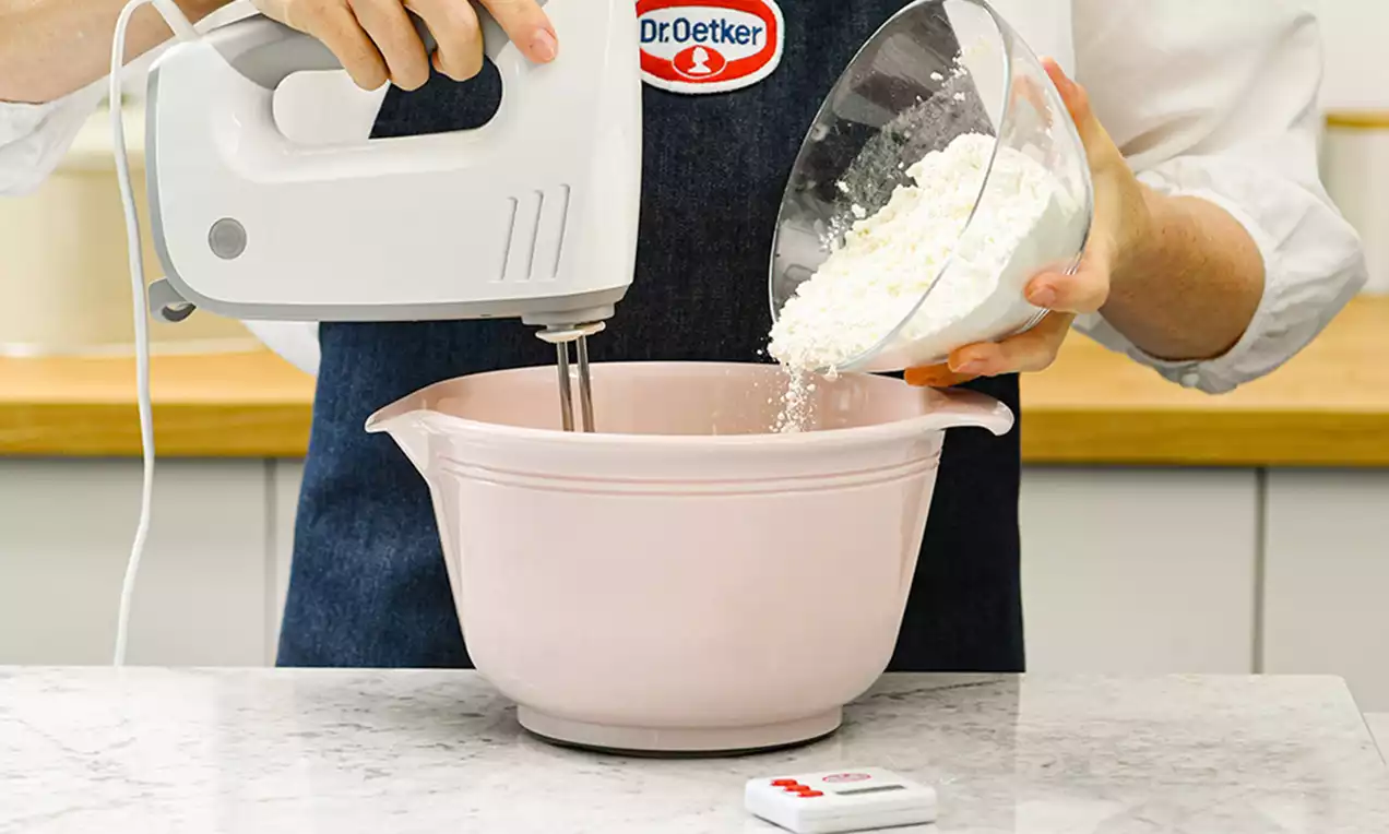 Pembe bir karıştırma kabında mikserle karıştırılırken beyaz un cam bir kaptan dikkatlice ekleniyor, fonda ahşap detaylı mutfak tezgahı görülüyor