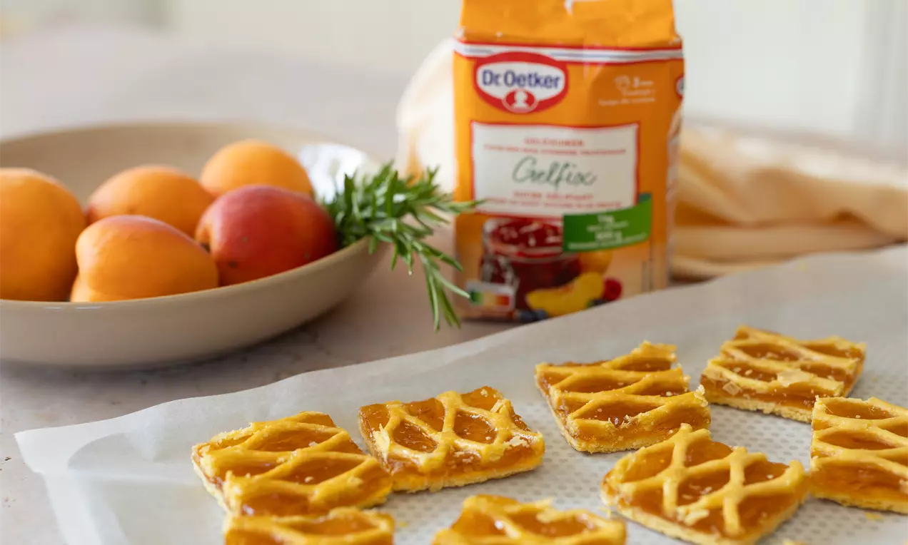 Vierkante koekjes met een gouden raster van deeg en glanzende abrikozenvulling op wit bakpapier, met een schaal abrikozen op de achtergrond