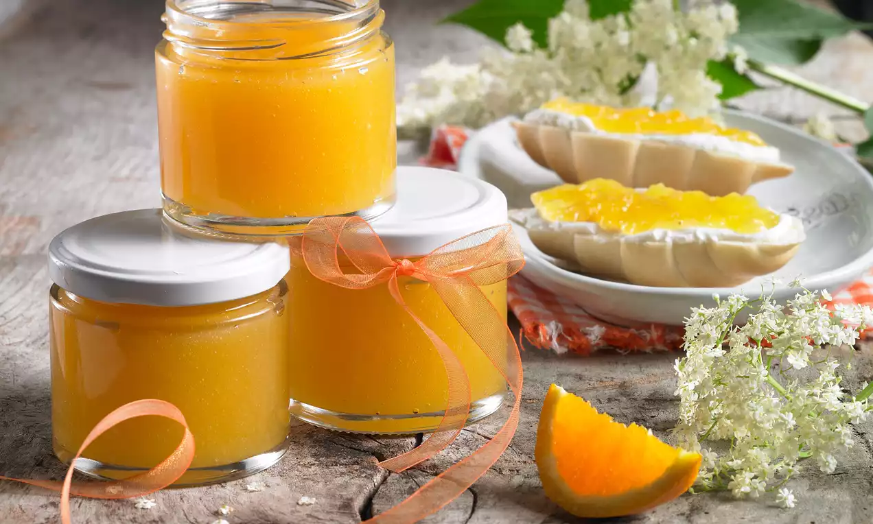 Drei Gläser mit leuchtend orangefarbenem Gelee, dekoriert mit Schleifen, daneben frische Holunderblüten und Marmeladenbrötchen auf rustikalem Holzbrett