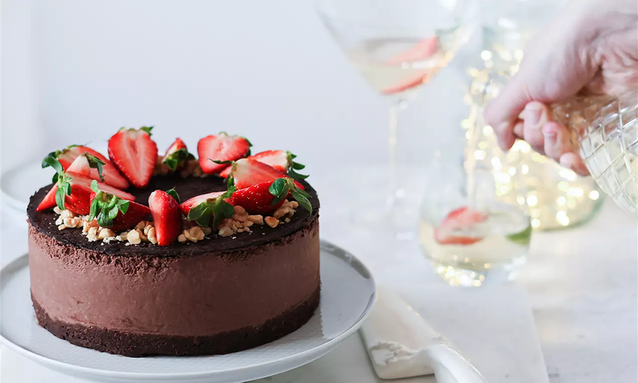 Кръгла сурова шоколадова торта с тъмнокафяв крем, гарнирана с резенчета ягоди и едро нарязани ядки на порцеланова чиния