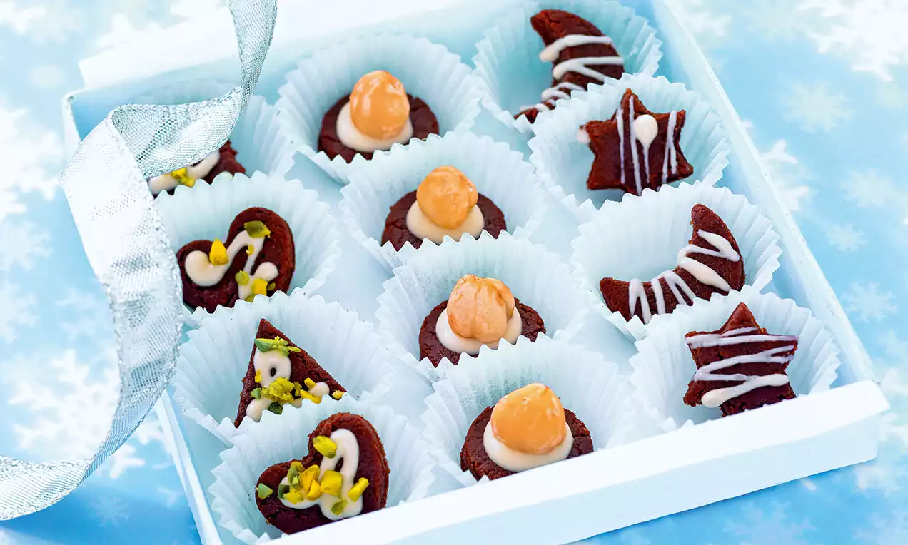 Petits biscuits au chocolat foncé décorés de glaçage blanc, pistaches et amandes, disposés dans des caissettes en papier bleu clair dans une boîte blanche.