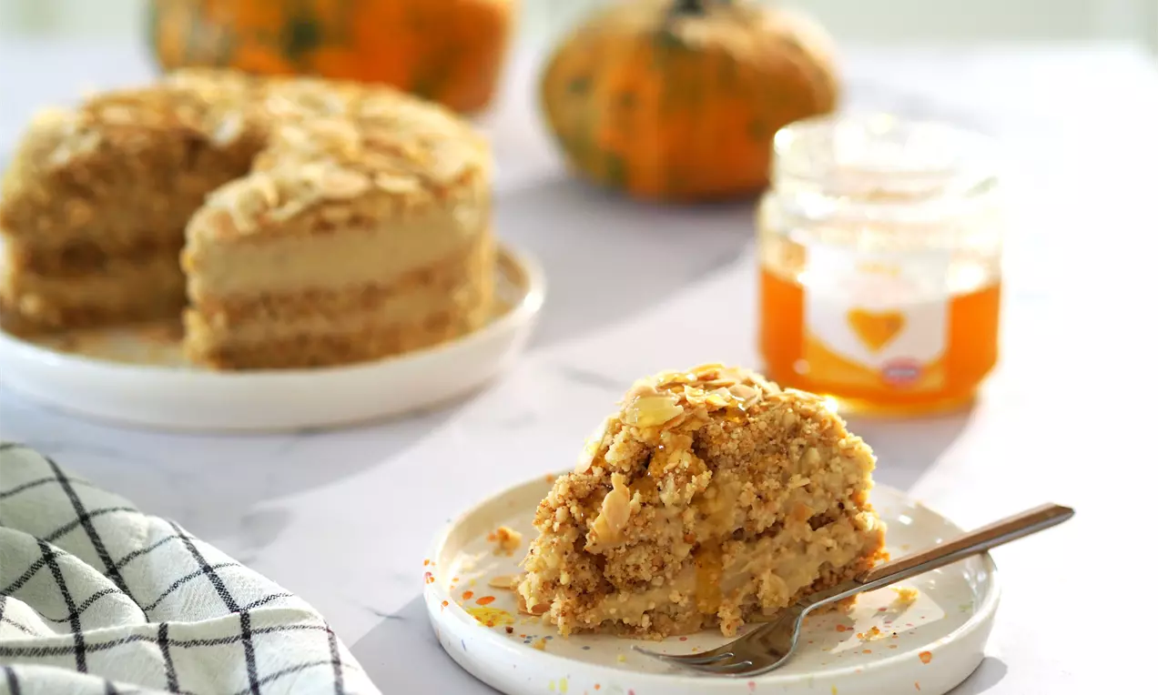 Светлокафяво парче медена торта с ронлива текстура и слоеве крем върху бяла чиния с вилица, заден план с буркан мед и тикви