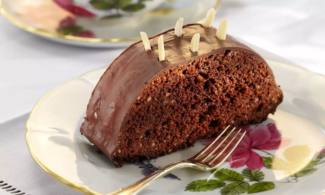 Halbkreisförmiger Schokokuchen mit glänzendem Überzug und gerösteten Mandelsplittern auf einem Teller mit floralem Muster und Gabel