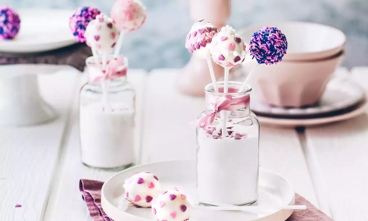 Cakepops med hvid chokolade dekoreret med lilla og lyserøde hjerter og perler, placeret på glas med hvidt fyld og bånd i lyserøde nuancer