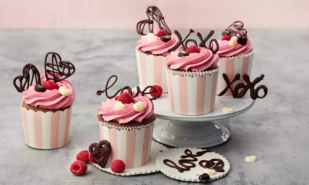 Schoko-Himbeer-Cupcakes mit rosa Creme, dekoriert mit Schokoschriftzug, Himbeeren und weißer Schoko auf gestreiften Förmchen und einer Tortenplatte