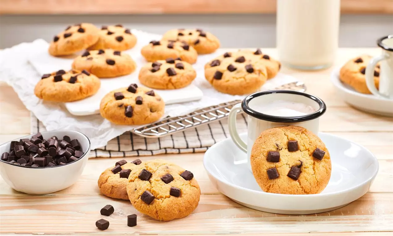 Zlatisté okrúhle cookies s jemne popraskaným povrchom, ozdobené kúskami tmavej čokolády, položené na papieri, tanieri a vedľa hrnčeka mlieka