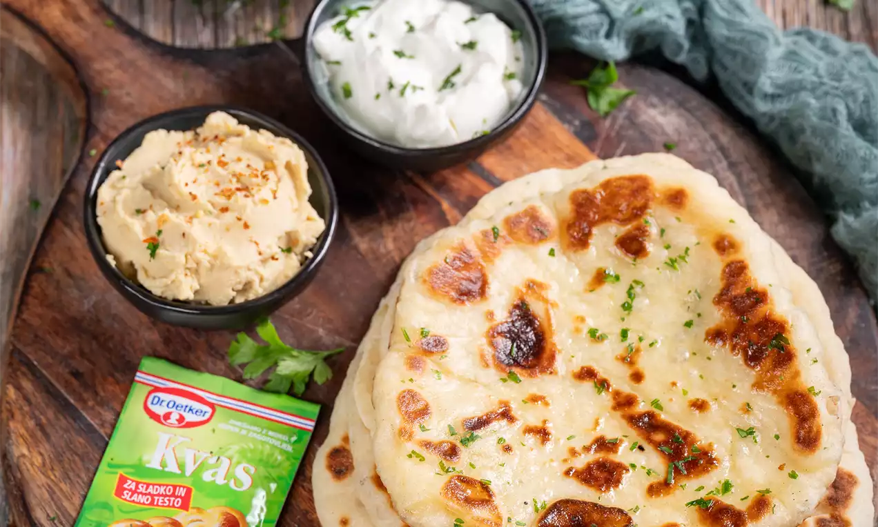 Zlato rjavi kruhki naan z mehko in rahlo papirnato teksturo, posuti z zelišči, naloženi na leseni deski ob skledicah omak