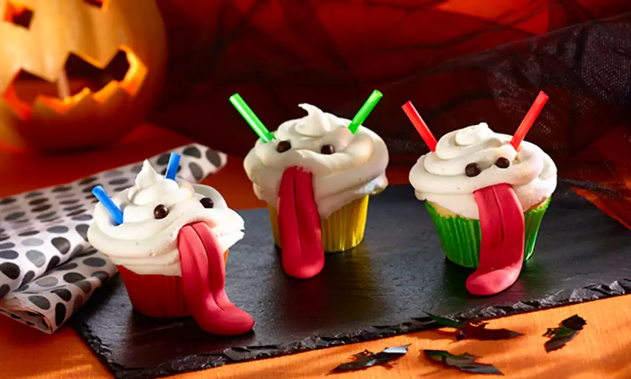 Halloween-cupcakes met witte toefjes glazuur, zwarte snoepoogjes, rode fondanttongen en gekleurde rietjes op een zwart bord