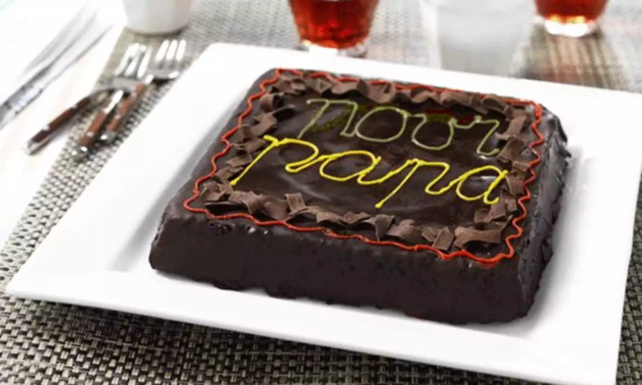 Gâteau carré au glaçage chocolat brillant garni de copeaux et d'une écriture jaune et rouge posé sur une assiette blanche