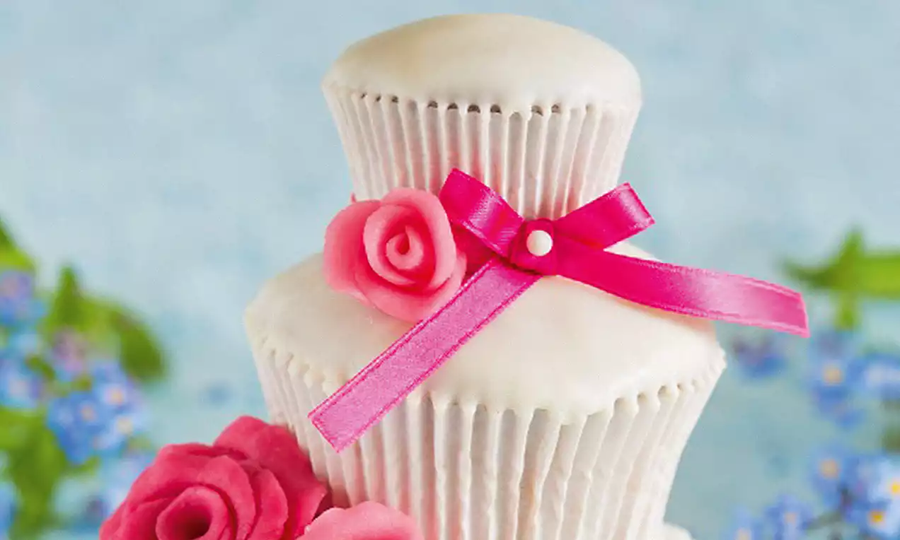 Mini-gâteau en forme de pièce montée à deux étages, glaçage blanc lisse, roses en pâte rose vif et ruban fuchsia sur fond fleuri