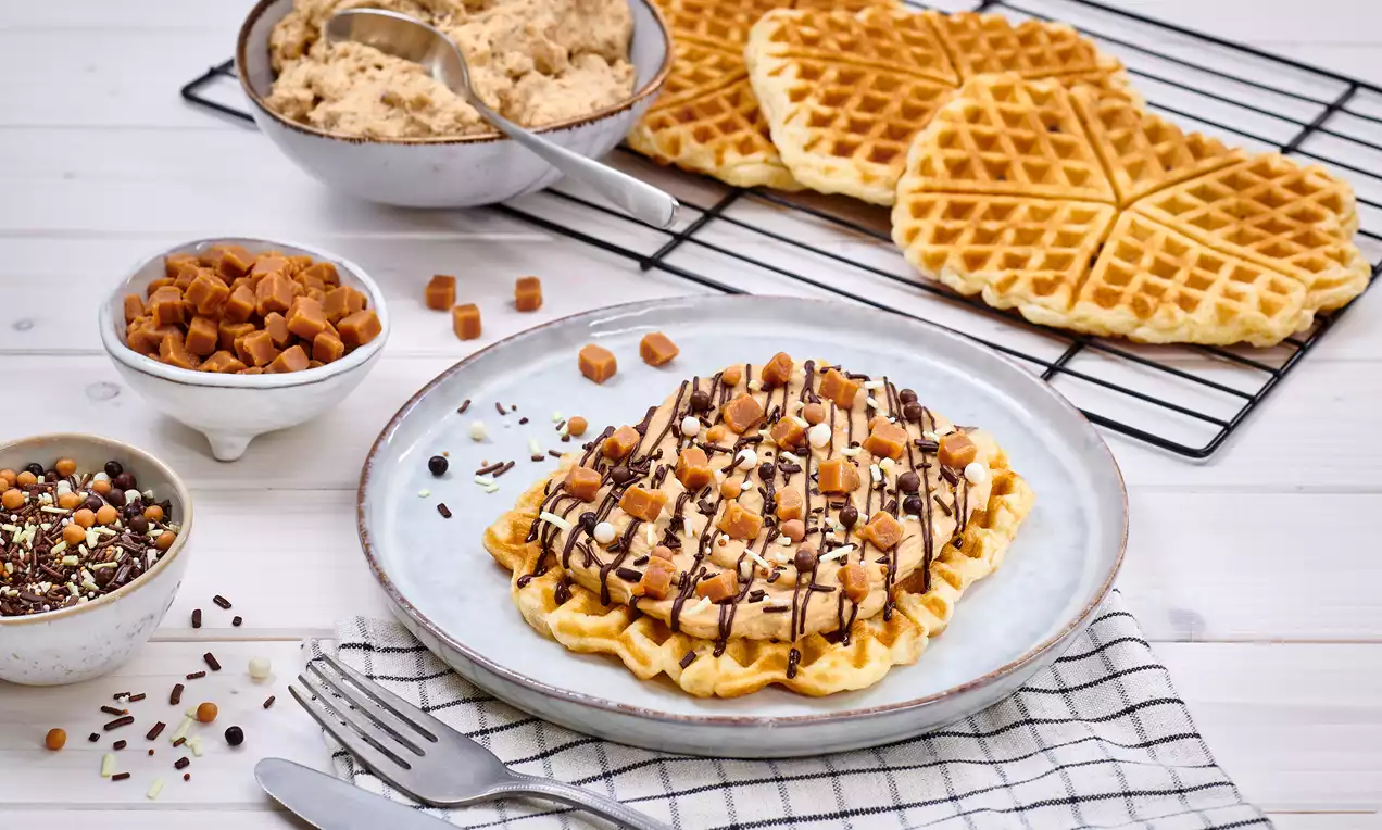 Goldbrauner, rechteckiger Croffle auf grauem Teller, dekoriert mit Schokosauce, Sahne, Streuseln und Karamellwürfeln, umgeben von Zutaten und Waffeln