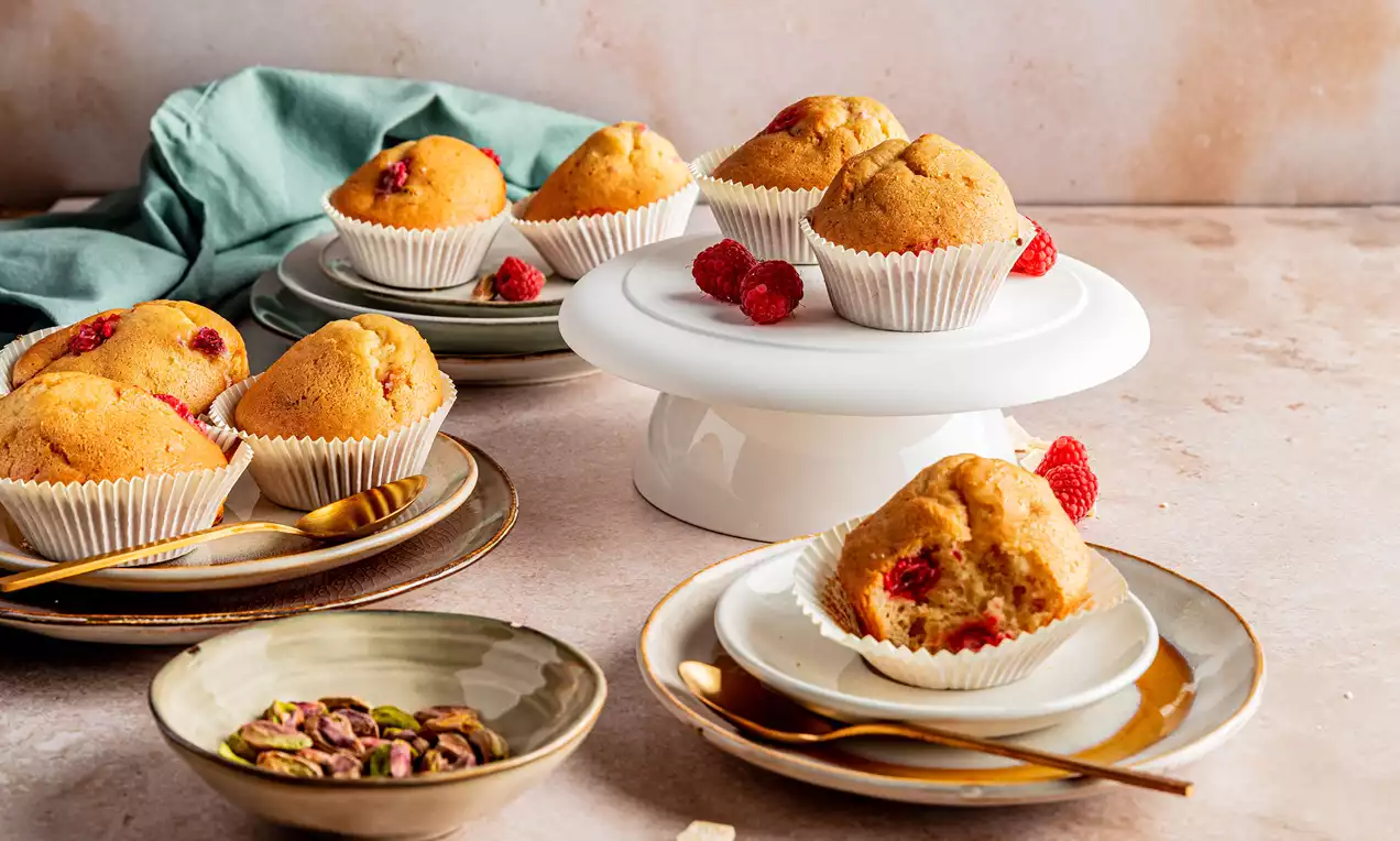 Goudbruin gebakken muffins met roze framboos en groene pistache, in witte papiertjes op borden en een witte schaal met losse decoratieve frambozen