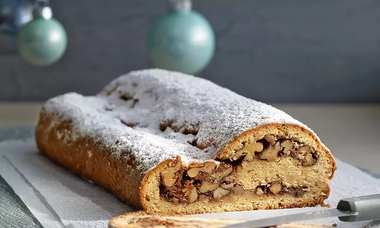 Mandel-Nuss-Stollen mit goldener Kruste und weißem Puderzucker, angeschnitten auf grauem Holzbrett, dekoriert mit schimmernden Christbaumkugeln