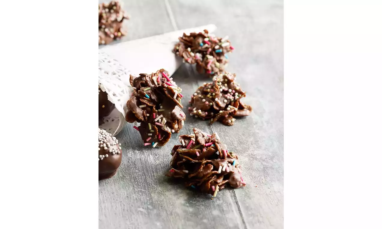 Sammenpressede hauger av mørk sjokoladeglasert cornflakes med sprinkles i farger og hvite sukkerperler på grått trebord