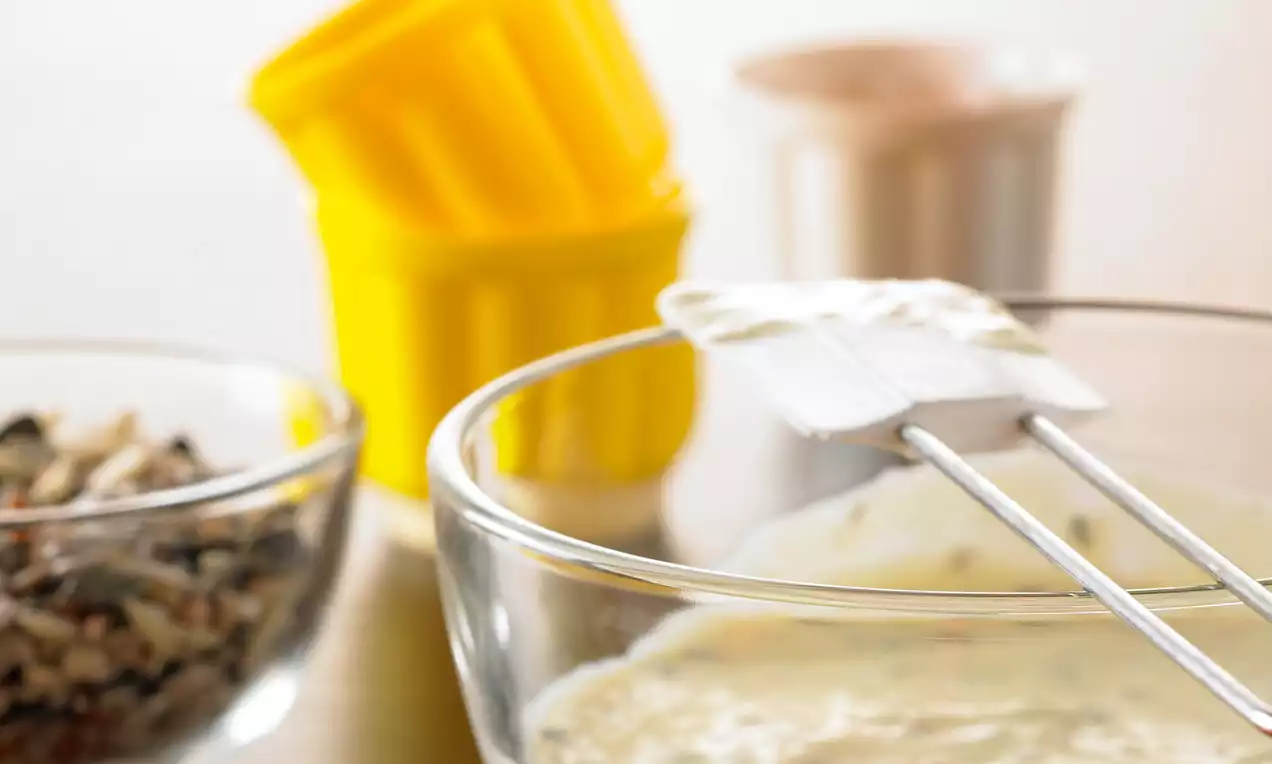 Crema bianca liscia in una ciotola di vetro, mescolata con una spatola, accanto a un recipiente di semi misti e stampi gialli