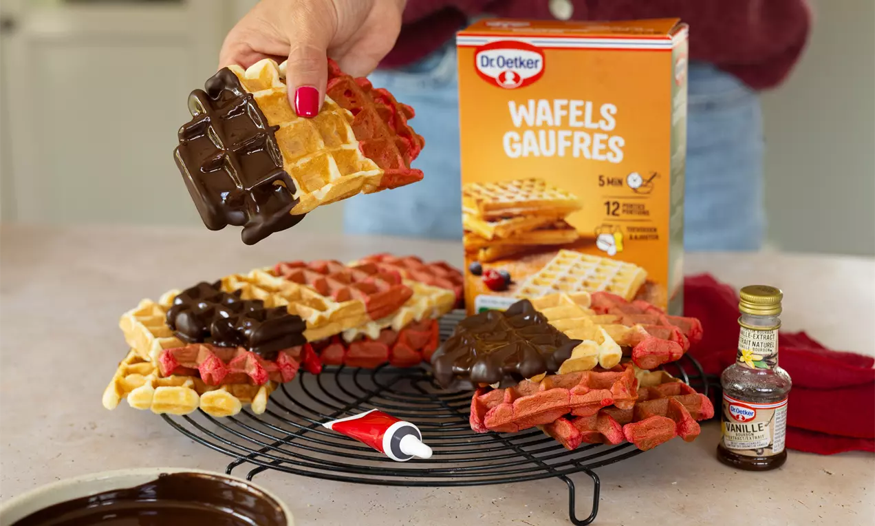 Gaufres dorées et rouge vif aux textures moelleuses, nappées de chocolat brillant sur une grille noire avec un tube de glaçage rouge