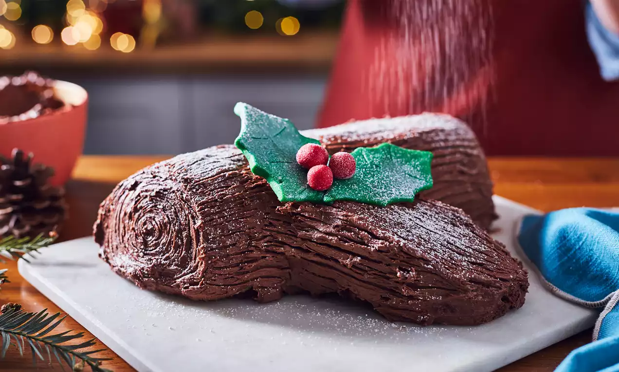 Chocolate Yule log with textured bark-like frosting, dusted with powdered sugar, topped with holly leaves and bright red berries on a marble board