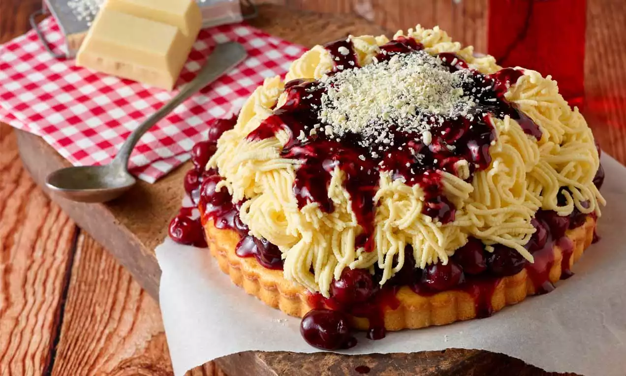 Spaghetti-Kuchen mit gelben Teighörnchen, roter Sauerkirschsoße und weißen Schokoraspeln auf Holzbrett mit kariertem Tuch und Besteck
