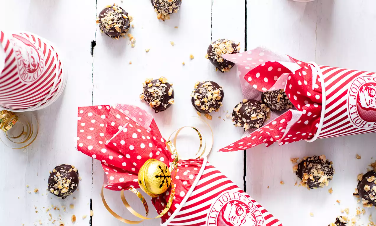 Kleine kugelrunde Marzipan-Pralinen mit dunkler Schokoladenhülle und gehackten Nüssen, verteilt auf weißem Holz neben rot-weißen Bonbonverpackungen