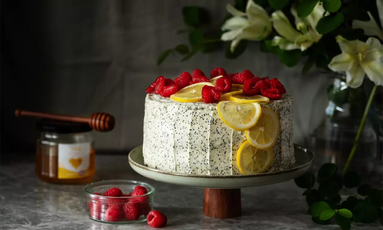 Кръгла торта с крем, осеяна с черни зрънца мак, украсена с резени лимон и свежи малини, поставена върху стойка на сив фон