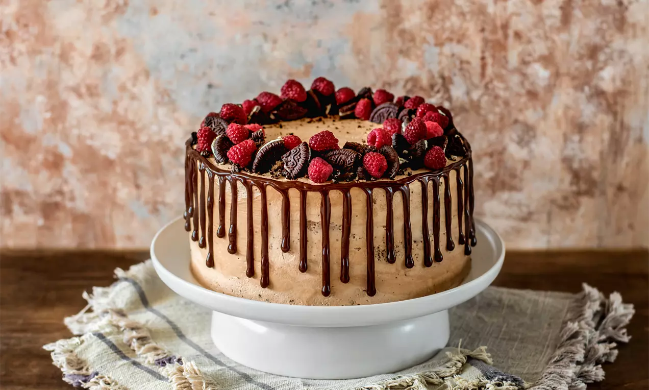 Кръгла шоколадова торта с млечнокафяв крем, течен шоколад, капещ по страните, гарнирана с пресни малини и бисквитки върху бяла стойка