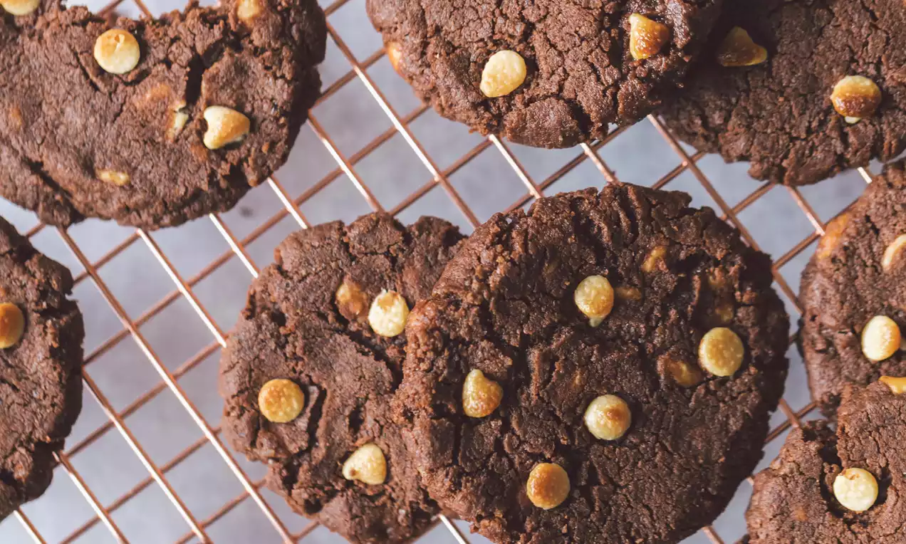 Galletas redondas de intenso color marrón oscuro y textura quebradiza, con gotas de chocolate blanco sobre rejilla de metal cobre brillante