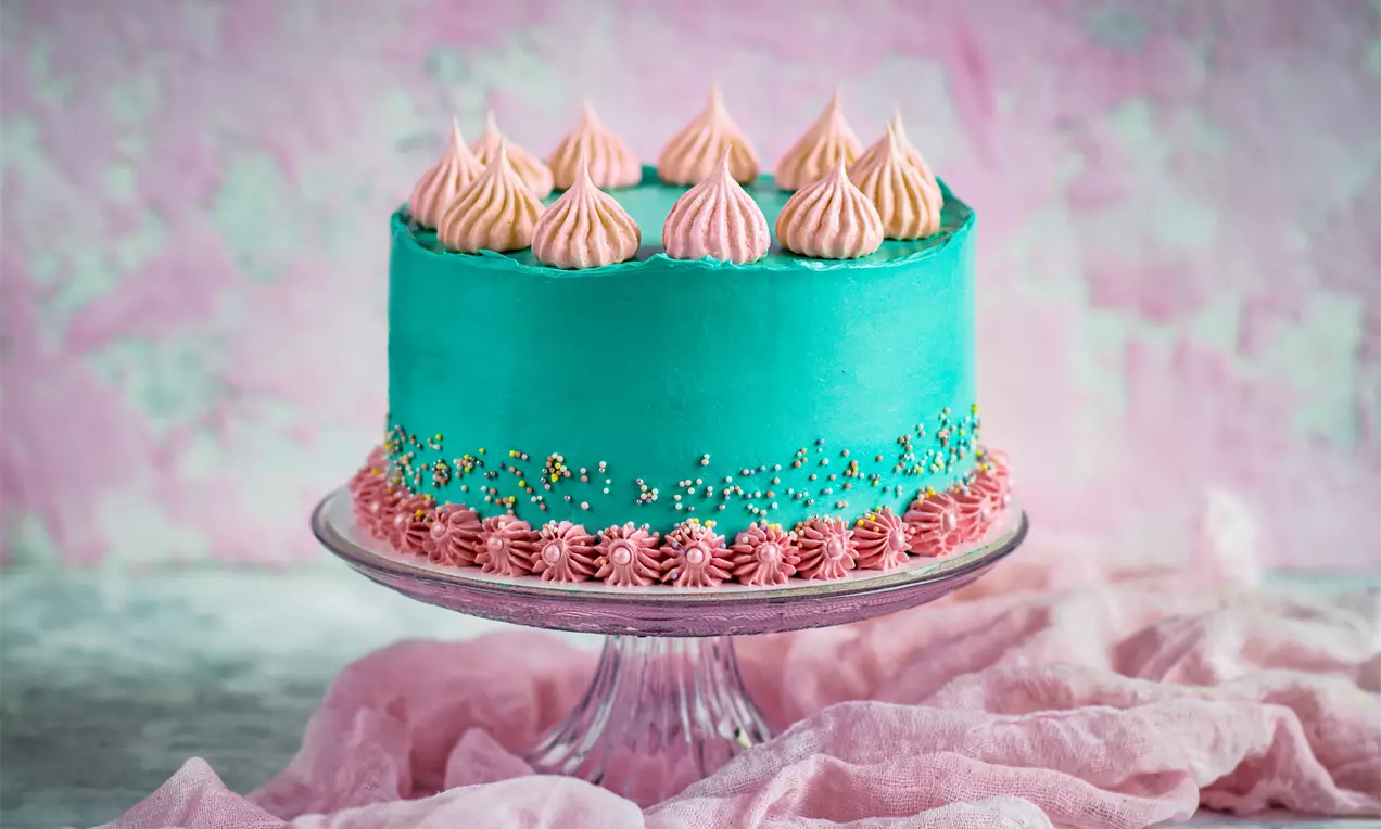 Тюркоазена кръгла торта с креместа текстура, украсена с розови звездообразни шапки от крем и поръска, поднесена върху стъклена стойка